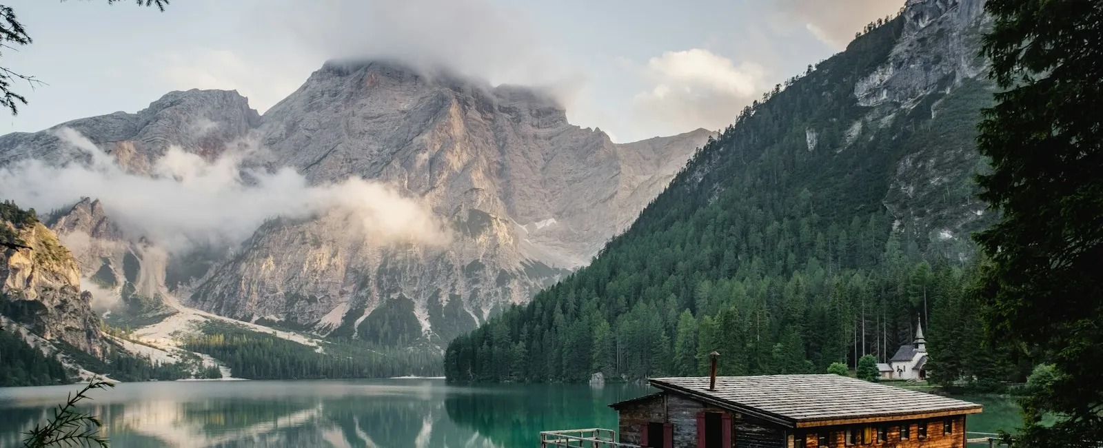 A lakeside cabin and still water beneath cloud-covered alpine mountains